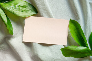Closeup of blank greeting, pink card and green large leaves on linen tablecloth background. Flat lay, top view. Spring, summer wedding, birthday, mother's day stationery mock-up scene.