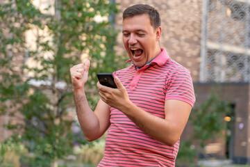 Naklejka na ściany i meble Excited screaming mature man using smartphone, celebrating mobile phone news and rejoicing while standing outdoor against building