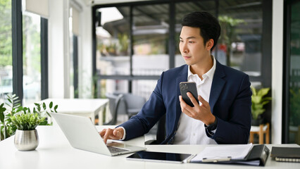Smart Asian businessman holding his smartphone and using laptop at his desk