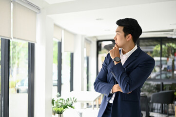 Thoughtful millennial Asian businessman hand on chin, looking out the window