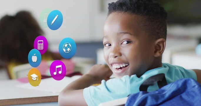 Animation of school icons over happy african american schoolboy sitting at desk in class