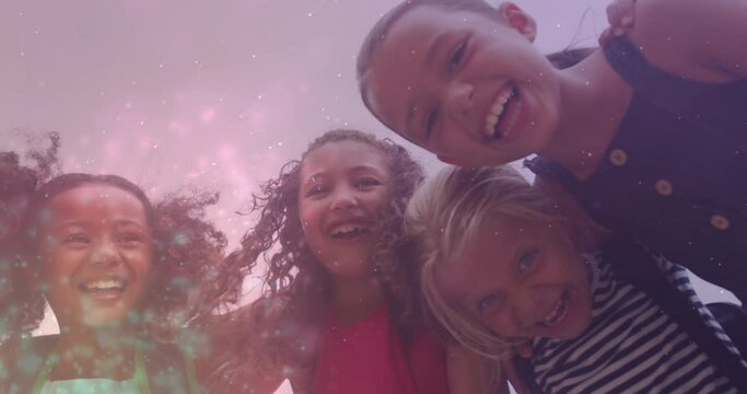 Animation of light particles over happy diverse schoolgirls embracing outdoors