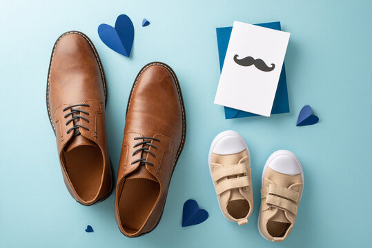 Little son shows love for dad on Father's Day. View from above of dad's leather shoes, baby boy's sneakers, heart embellishments, and postcard with mustaches on pastel blue background