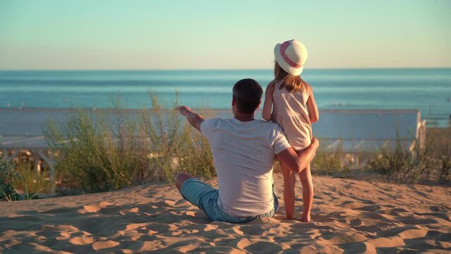 Back view of father and child look at hotel beach with blue sea at sunset. Concept of summer vacation, travel, holiday. People outdoors on seascape