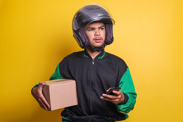 Asian online taxi driver motorbike wearing green jacket and helmet while holding smartphone and package isolated on yellow background