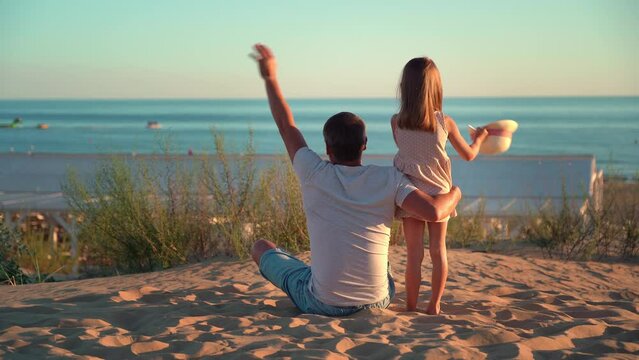 Back view of father and little daughter on beach wave hands boat sailing on blue sea at sunset. Concept of summer vacation, travel, holiday. People outdoors on seascape