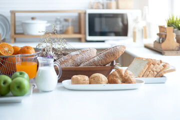Variety of fresh fruits and bread on table in bright white kitchen, healthy lifestyle concept, glass of milk and orange Juice