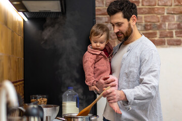 Father cooking meal holding toddler daughter in kitchenn.