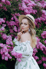 Beautiful, stylish, cute blonde in a white dress with a green pattern as a model for a photo shoot in lilac. They made luxurious portraits of her in the green garden of the park.