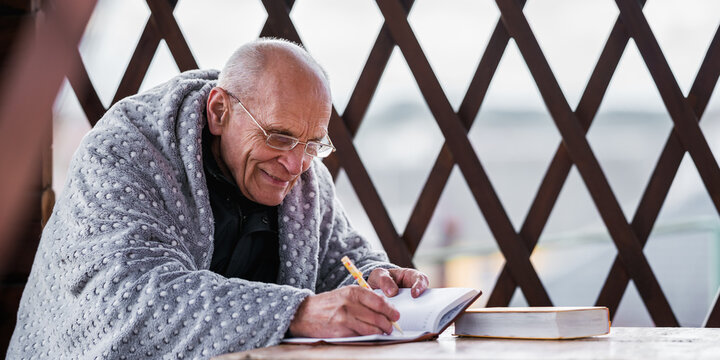 Positive elderly person in glasses planning daily chores seated at table with notepad and pen in hand in cottage alcove outdoor