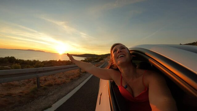 PORTRAIT, LENS FLARE: A cute lady smiles through window of a car driving by sea. She is enjoying breeze in her hair and freedom while driving to her summer vacation on beautiful coast of Adriatic Sea.