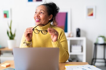 Pensive elderly Ethiopian female employee with eyeglasses looking away and smiling while analyzing business plan and working remotely with portable computer