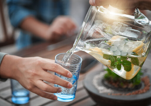 Hands, closeup and water into a glass, outdoor and social gathering with friends, drink and cool down. Zoom, people and friends with clear liquid, ice and lunch with fresh mint, lemon and natural