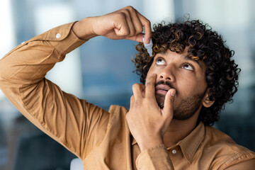 A young businessman works for a long time at a laptop inside the office, the man drips drops into...