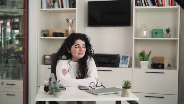 The young brunette woman with long curly hair sits at the desk in the office; she is very sad and worried about her work