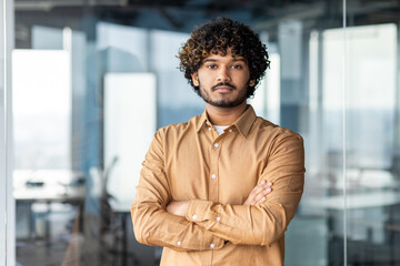 Portrait of a successful young hispanic programmer standing in a modern office with his arms...