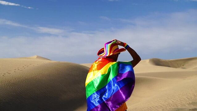 Gay boy proudly displaying rainbow flag outdoors in beautiful setting with sand dunes, ocean, seagulls and blue sky of Maspalomas, Canary Islands