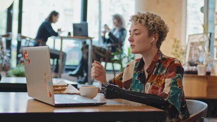 Modern young woman with disability wearing arm prosthesis sitting at cafe table talking on video call on laptop - Powered by Adobe