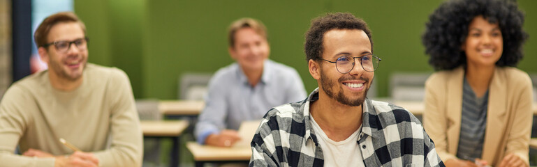 Young cheerful mixed race male office worker sitting listening to coach during seminar or training and smiling while studying with colleagues in coworking space