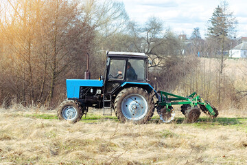 Obraz premium Blue small modern tractor machinery plowing agricultural field meadow at farm on sunny day. Farmer cultivating and make soil tillage before seeding plants and crops, nature countryside rural scene
