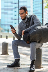 Young african american man sitting outside using mobile phone with travel bag