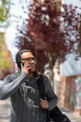 Smiling young african guy with bag walking outside and talking on mobile phone