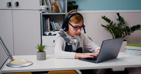 Joyful little boy in eyeglasses and headphones is playing video game using laptop sitting at his desk in living room. Gamers lifestyle.