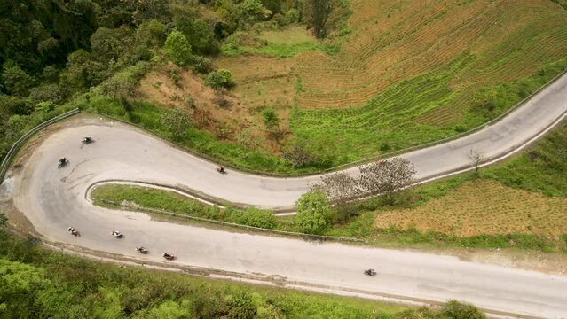 Ha Giang Loop, Vietnam. Aerial drone shot of motorbikes on dramatic winding road down mountain, tilting up to reveal road disappearing across valley and up into distant misty peaks on horizon.