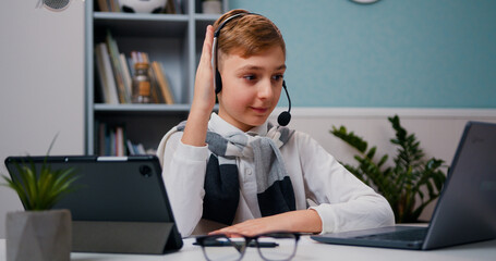 Closeup view of cute boy raises his hand and answers. Kid pupil in headphones attending to home online school class on his computer laptop.