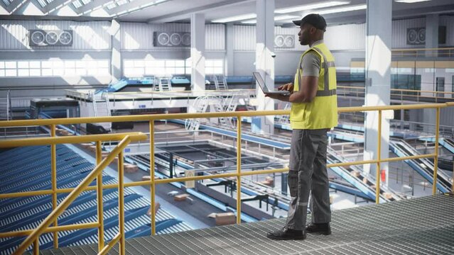 African American Man Working in a Modern Advanced Logistics Center with Automated Belt Conveyors for Sorting Orders. Black Male in High Visibility Vest Using Laptop Computer in an Industrial Facility