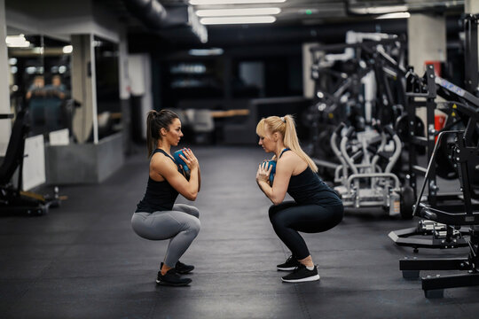 Two muscular female friends are squatting and holding kettlebells during their training in a gym.