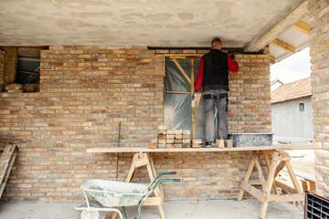A housebuilder is standing on a scaffolding and stacking bricks on a brick wall.