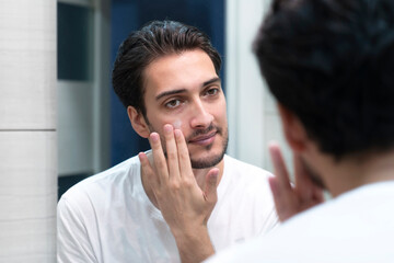 Fototapeta na wymiar Handsome man looking at mirror and applying moisturizing cream on cheeks in bathroom. Groomed young guy doing skincare morning routine after taking a shower