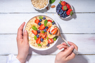 Bowl of sweet breakfast oatmeal with berry. Portion plate of wholegrain oats porridge, with yogurt, fresh banana, strawberry, blueberry, almond nuts, on white wooden table. Healthy summer morning food