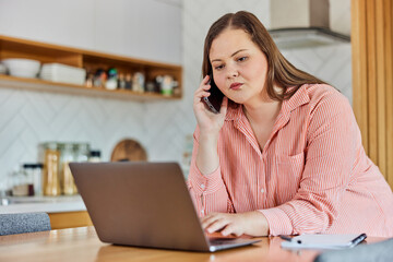 Fototapeta na wymiar Plus-size businesswoman using a laptop while talking on a mobile phone at a desk at the kitchen table.