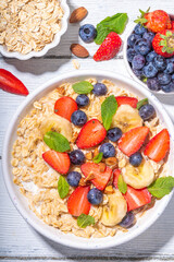 Bowl of sweet breakfast oatmeal with berry. Portion plate of wholegrain oats porridge, with yogurt, fresh banana, strawberry, blueberry, almond nuts, on white wooden table. Healthy summer morning food