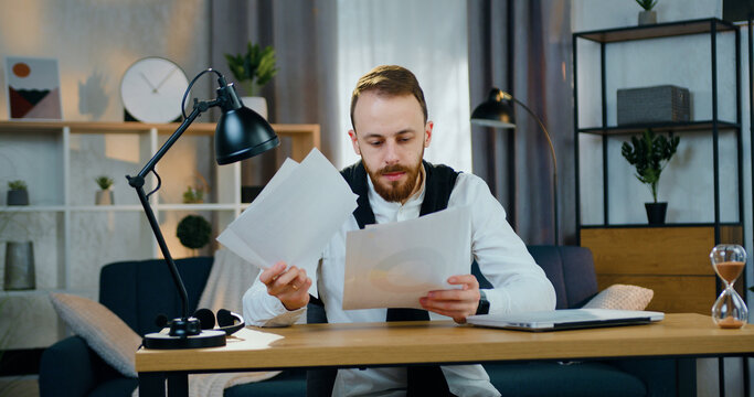 Handsome confident concentrated young bearded man sitting at his workplace at home and revisioning different reports during remote work from home,close up