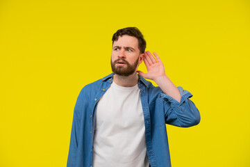 Young, serious, blond man in a blue shirt on a yellow background, holding an ear to listen better,...