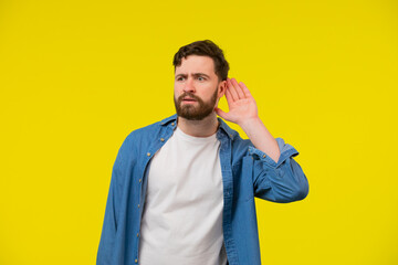 Young, serious, blond man in a blue shirt on a yellow background, holding an ear to listen better,...