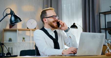 Handsome cheerful young bearded guy in glasses laughing from joke that hearing from his interlocutor on phone during working on laptop at home