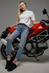 Fototapeta na wymiar Studio shot of trendy blond haired woman and her powerful modern motorbike against gray background.