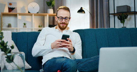 Likable smiling happy young bearded man in glasses sitting in relaxed pose on soft couch at home and browsing phone apps ,leisure concept
