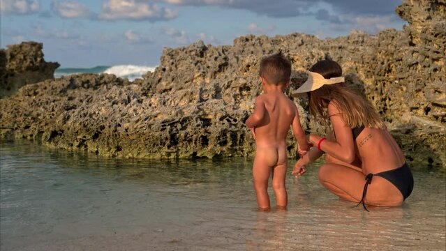 Cute young tanned latin baby boy toddler with his brunette mother watching the water at a rocky beach searching on a warm sunny afternoon