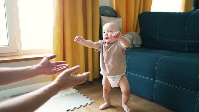 First steps.Happy child takes his first steps under supervision of his father. Parental helping hand. Father's Day. Toddler at home walks on floor of laminate. First steps are joy of child and toddler