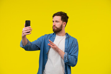 Happy young man video calling, talking online with mobile phone, saying hello to smartphone camera and waving hand friendly, standing over yellow background