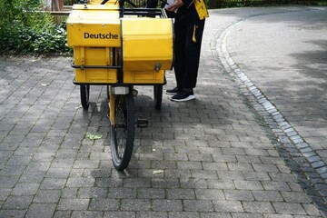 In schwarz-gelbgekleideter junger Postbote steht vor gelbem Fahrrad der Deutschen Post auf Gehweg...