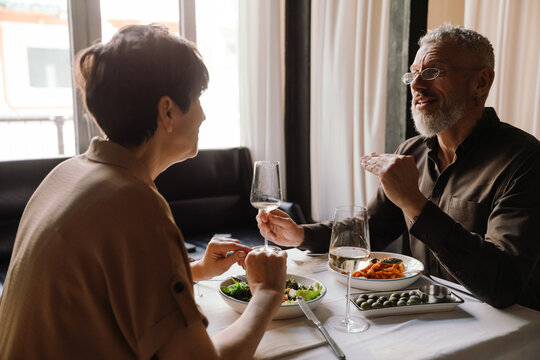 Positive mature couple dining with glasses of wine and talking in cafe