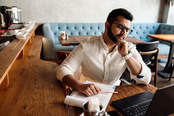 Sad busy indian male business owner in eyeglasses filling orders on laptop in cafe