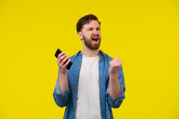 Overjoyed happy tanned handsome man in blue basic t-shirt raise fist up using phone posing isolated on orange yellow studio background. Copy space Banner Mockup. Online Social media. Lucky win concept