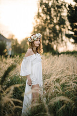 A beautiful tender young girl in a white dress with a wreath of flowers on her head is standing in a spring field at sunset. Happy bride with blonde curly hair enjoying sunny day. Summer tender photo.